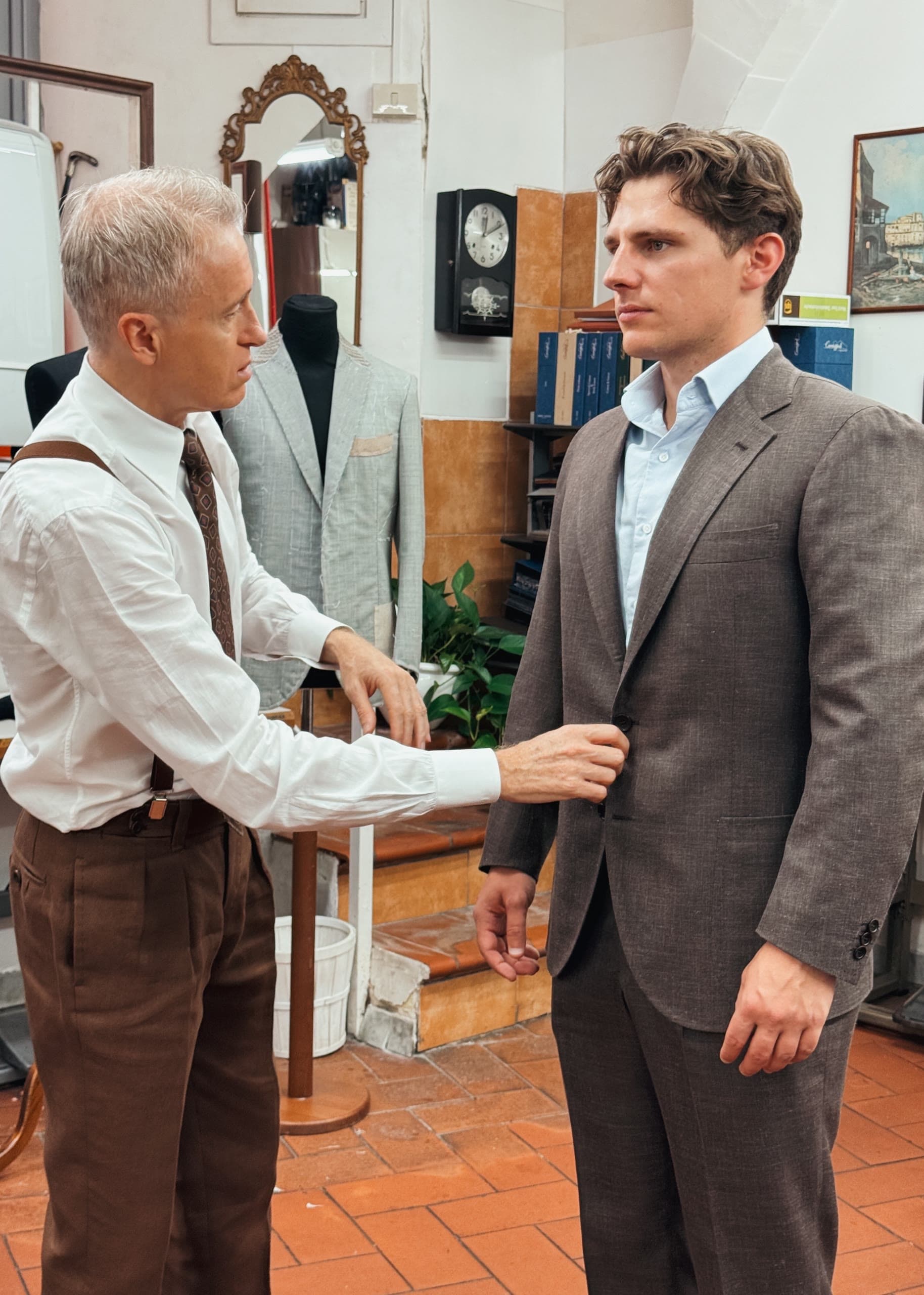 A tailor adjusts the front closure of a brown suit on a client during a fitting.