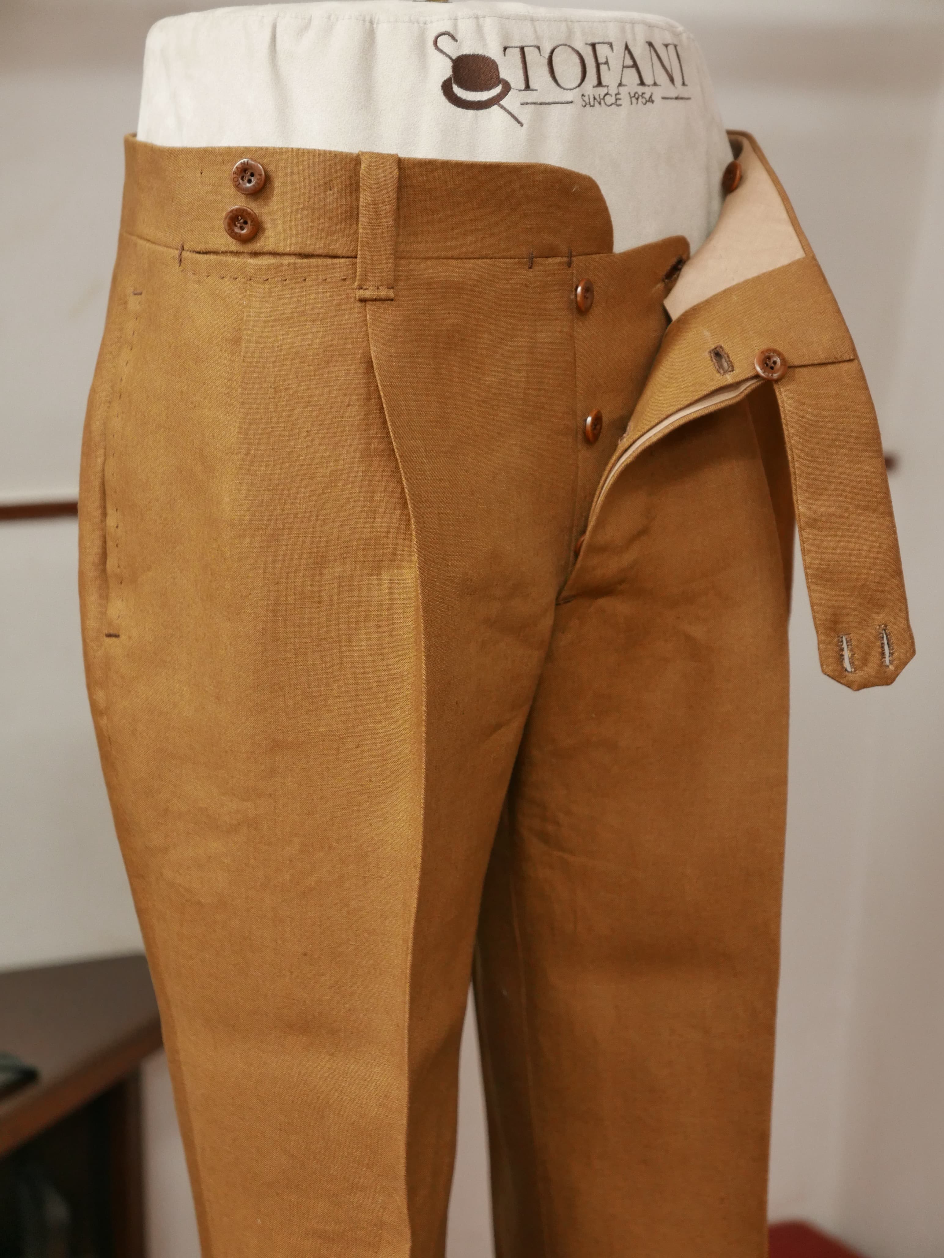 Close view of beige bespoke trousers on a form, showing the waistband, button fly and pleat.