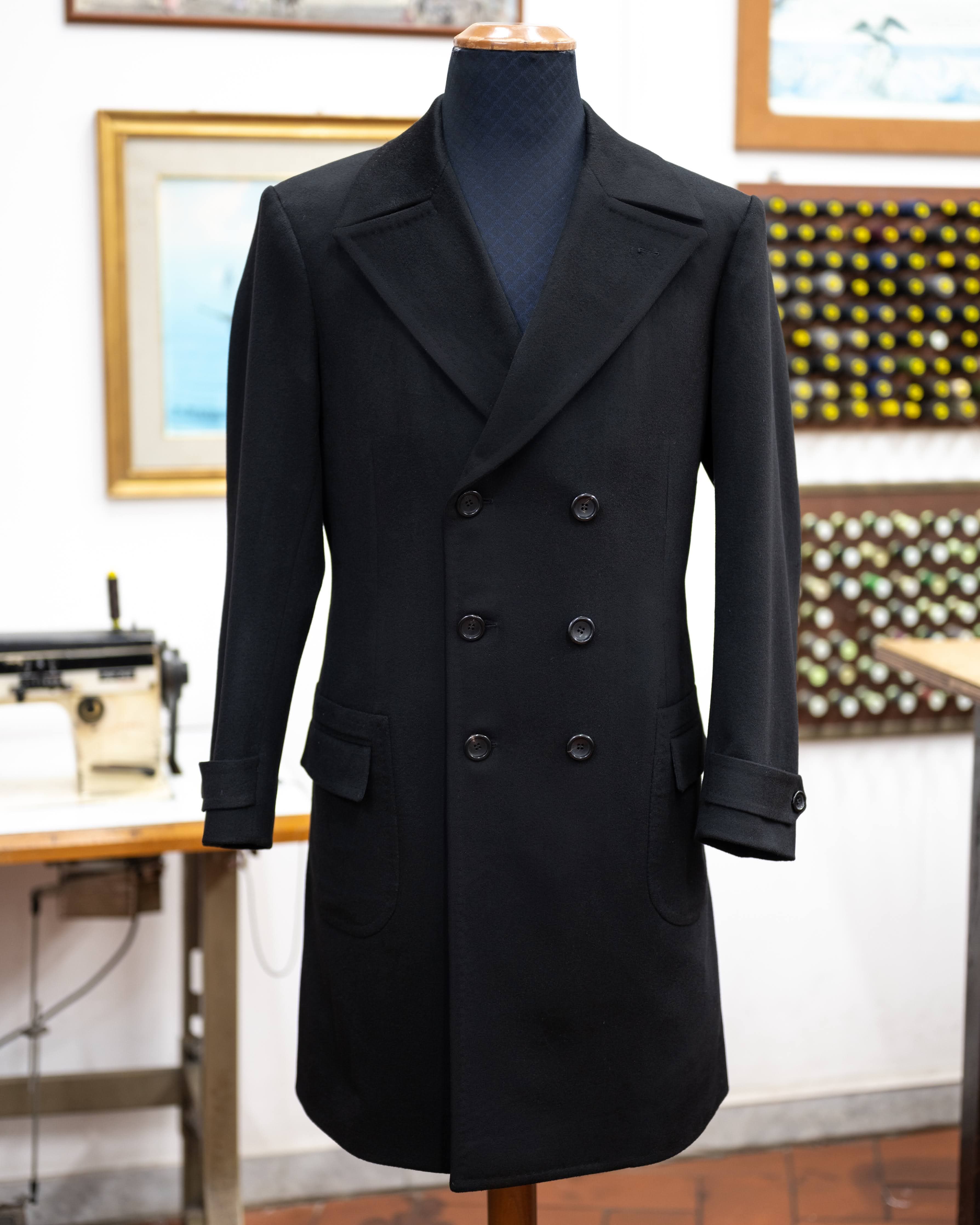 A black double-breasted bespoke overcoat with peak lapels, displayed on a tailor’s form.