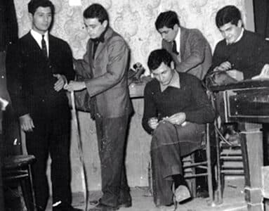 Black-and-white photograph of a tailoring workshop: one man is fitted for a jacket while others sew by hand.