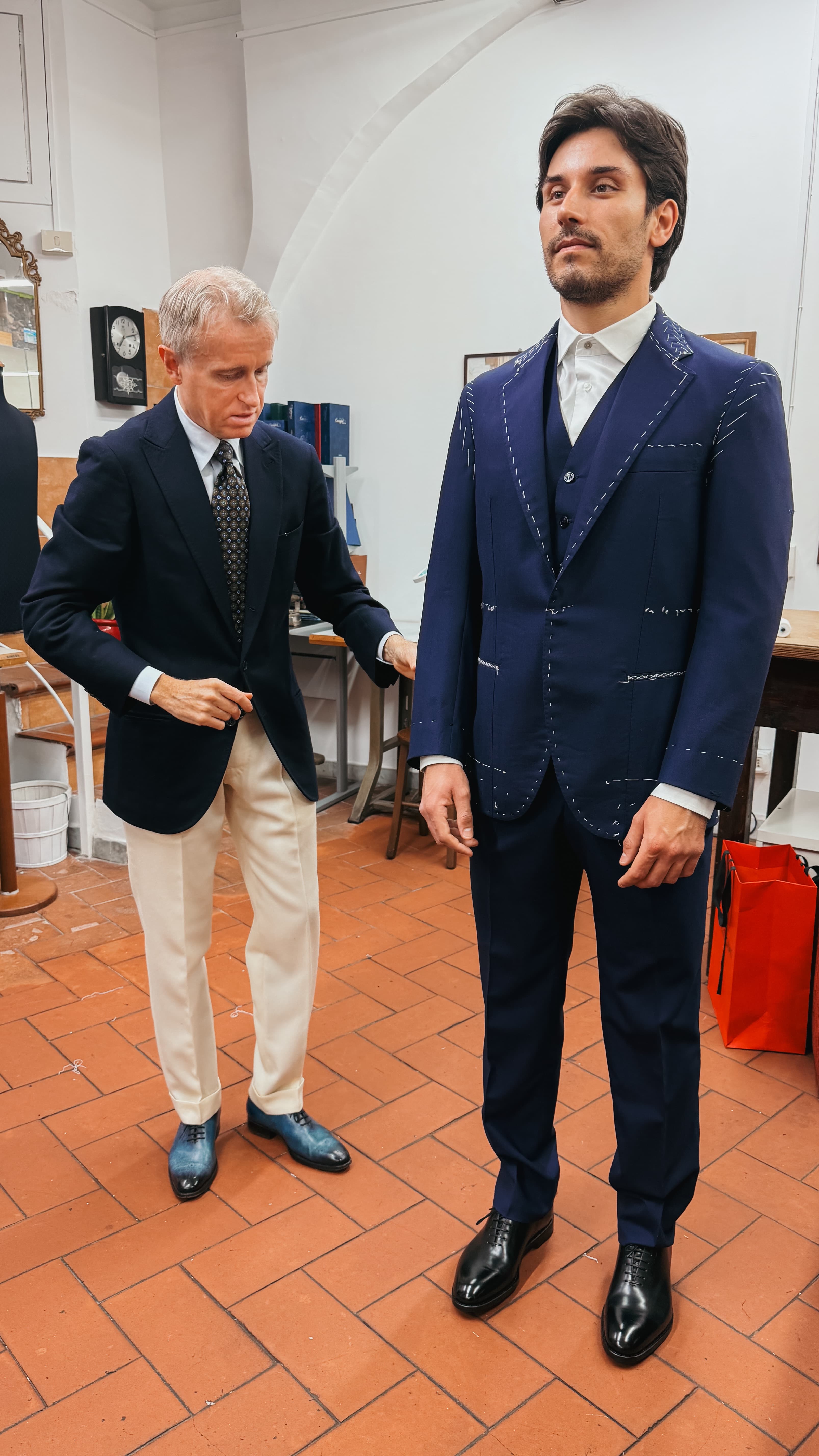 A tailor adjusting the sleeve of a navy suit during a fitting, on a terracotta tiled floor.