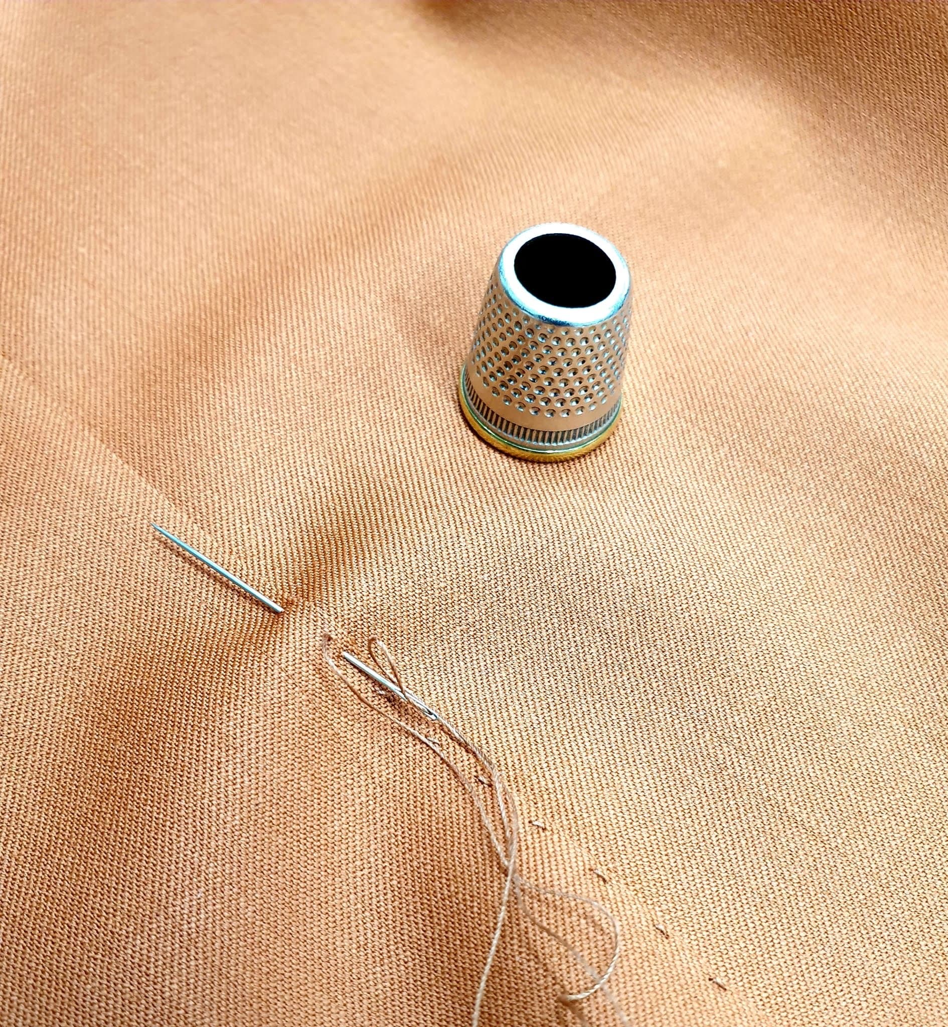A needle and a silver thimble on tan cloth, mid-stitch.