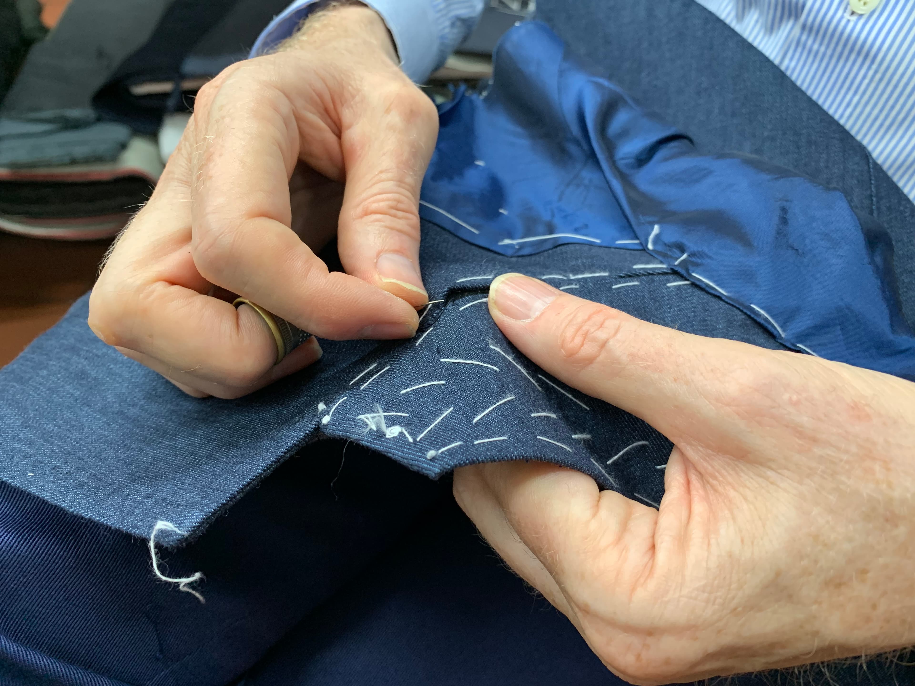 Hands sewing a navy jacket, with white basting thread and a metal thimble.
