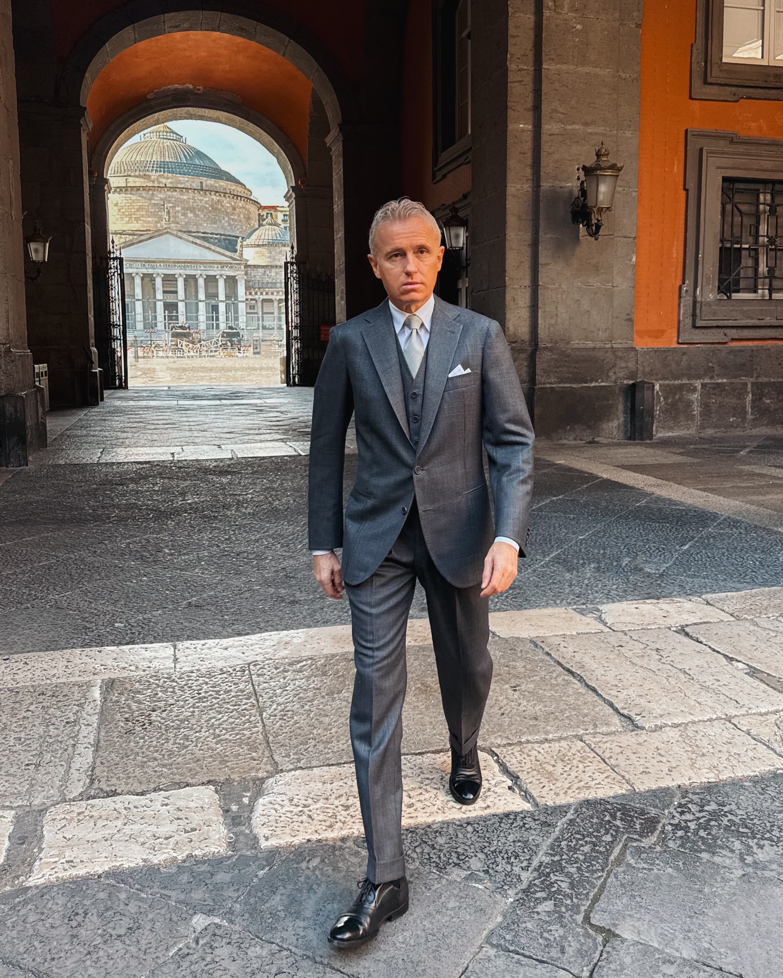 A man in a charcoal three-piece suit walking through a stone archway in Naples, with a colonnaded square beyond.