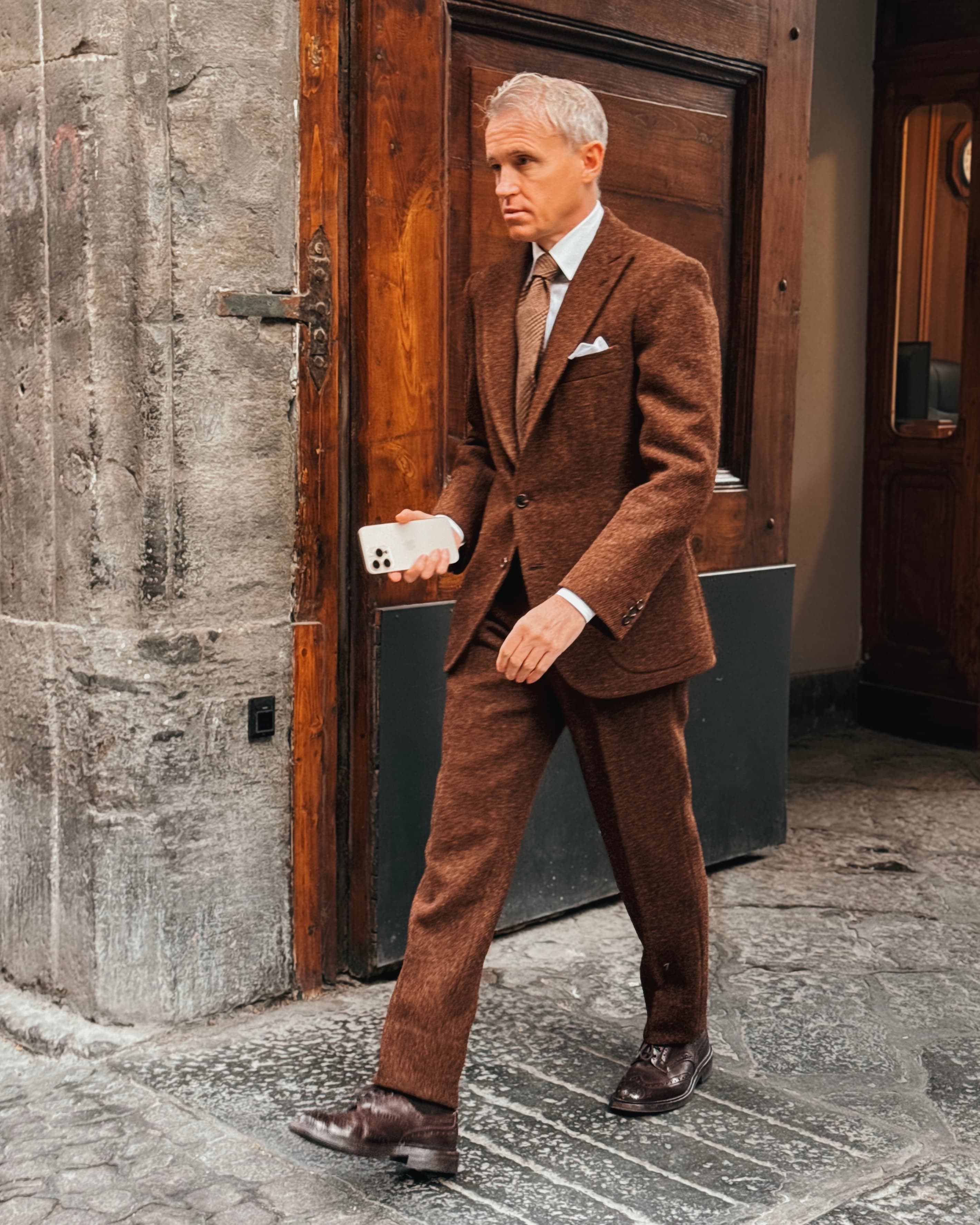 A man in a tobacco-brown suit walking past a weathered wooden door.